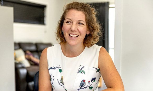 Headshot of woman smiling, wearing white sleeveless dress, leaning on a table